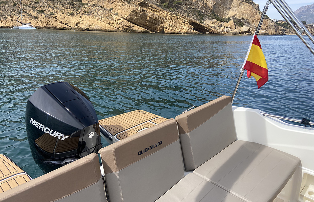 Quicksilver 675 Sundeck con cómodos asientos ergonómicos en popa, bandera española y acantilados de la costa de Albir en Alicante al fondo