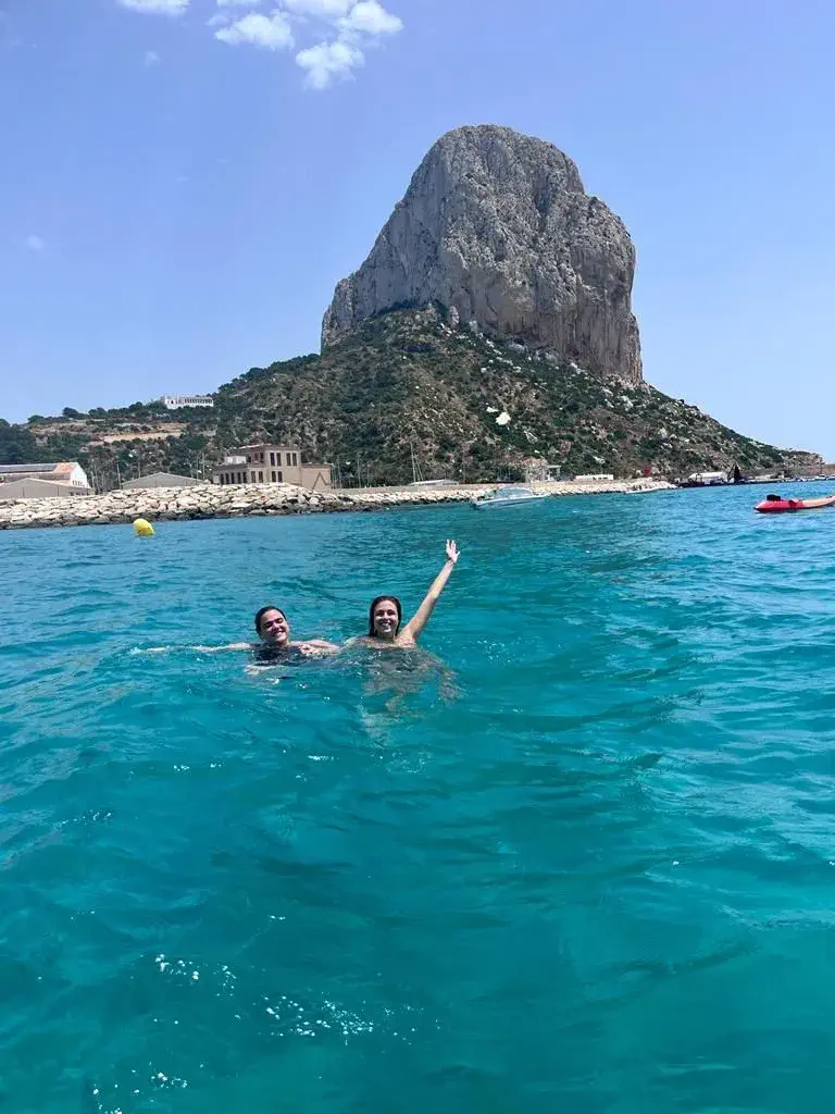 Two friends swimming and waving in the water with the Pe&ntilde;&oacute;n de Ifach in the background on the coast of Calpe